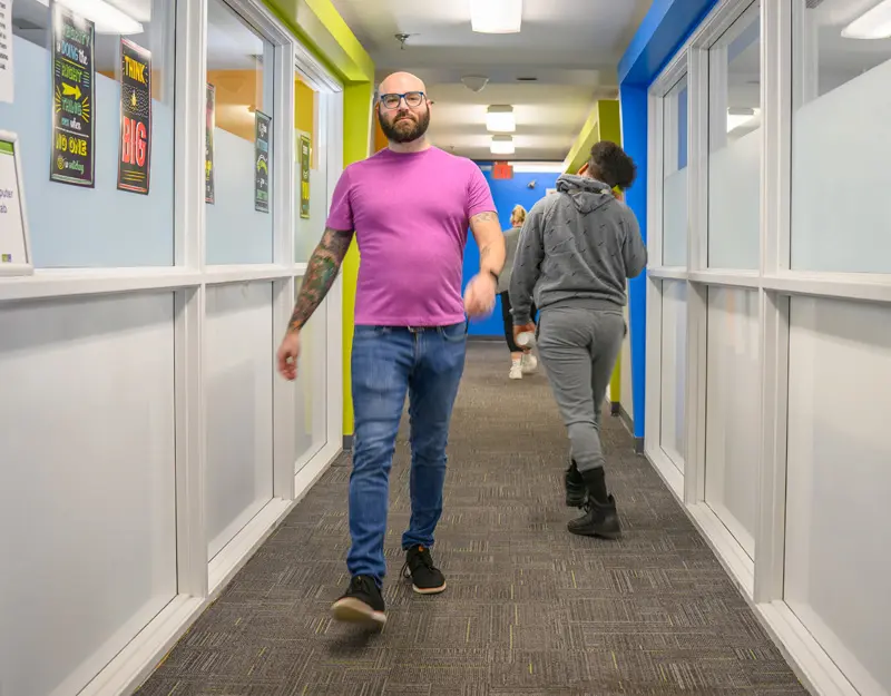 Phil Dejean, in a pink shirt, walks down a hallway.