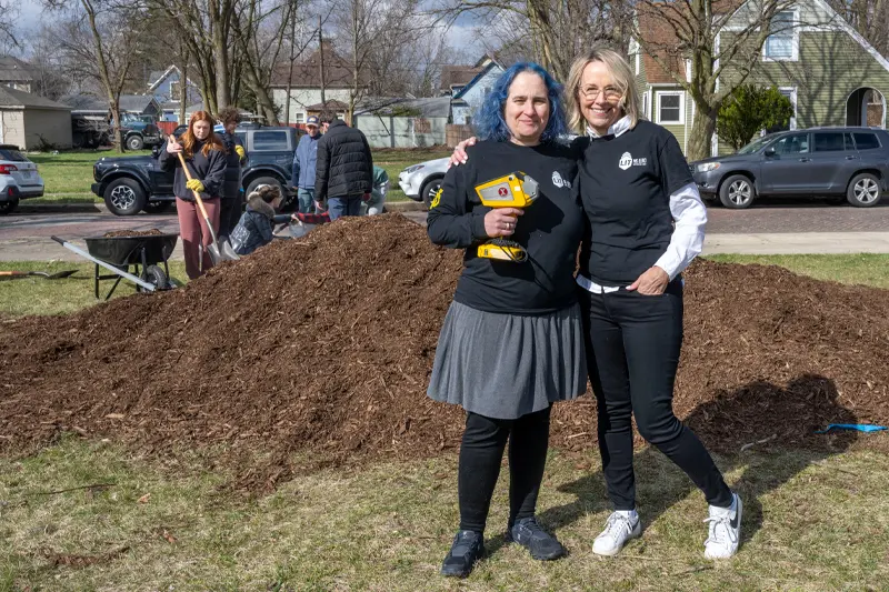Marya Lieberman and Heidi Beidinger, co-founder of ND-LIT stand In front of a mulch pile for the Mulch Madness event in South Bend. Marya is holding a yellow lead-testing device in her hand.