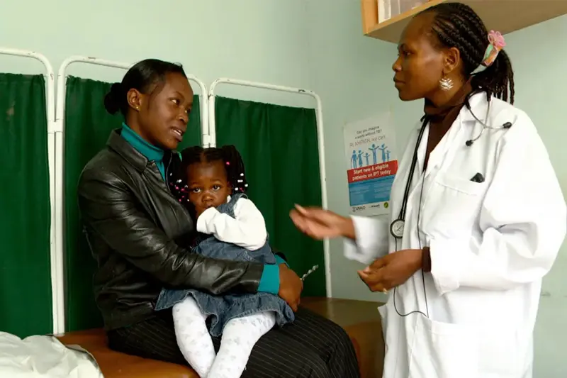 A black woman and her toddler daughter sit on an examination table talking to a black doctor.