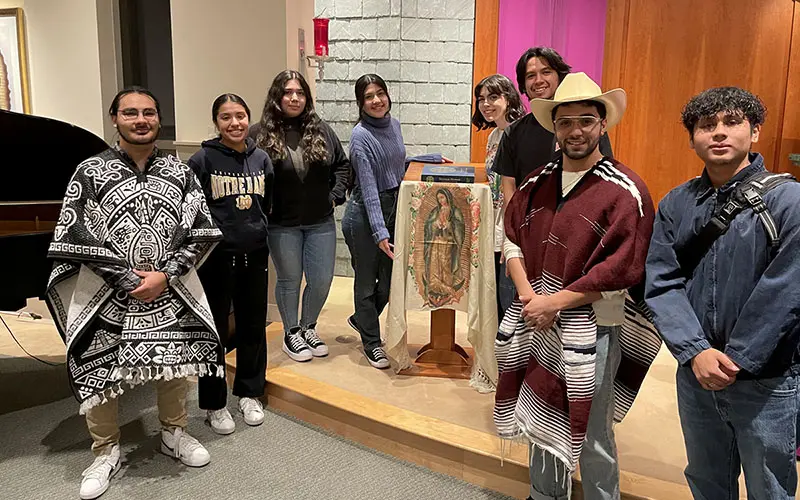 Seven students stand beside a small table draped with a cloth featuring an image of Our Lady of Guadalupe.  A hymnal rests on top of the table. Two students wear patterned ponchos. One student wears a Notre Dame sweatshirt. The students are gathered in a room with gray stone walls and a purple curtain. A portion of a piano is visible on the left side of the image.