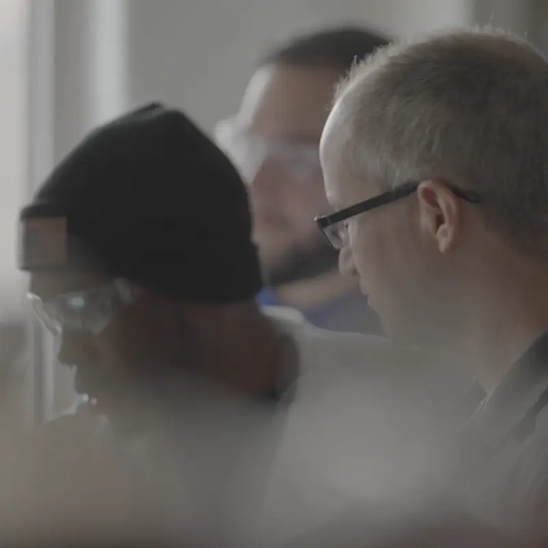 Three individuals, two wearing safety glasses and one with a black knit hat, are engaged in a research activity in a lab setting.